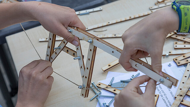 Kinder entwerfen und bauen eigene Brückenmodelle. Mit gelochten Holzteilen, Schrauben und Schnur entstehen kleine Konstruktionen, bei denen ausprobiert wird, wie sich stabile Strukturen mit möglichst wenig Material realisieren lassen.
